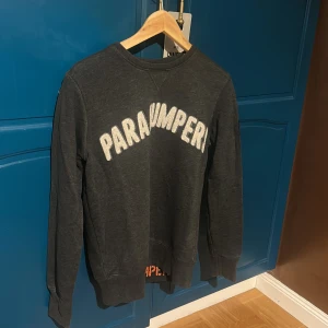 Mörkgrå fleece-tröja Parajumpers  - Snygg mörkgrå fleece-tröja från Parajumpers med broderad logga på ärmen och stor vit text på bröstet. Tröjan har lång ärm, rund hals och en loose passform. Perfekt för dig som gillar streetwear och vill ha något bekvämt och stilrent.