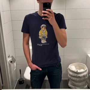 Svart Polo Bear t-shirt från Ralph Lauren - Mörkblå t-shirt från Polo Ralph Lauren med ikoniskt Polo Bear-tryck på bröstet. Björnen har gul jacka och keps, vilket ger en cool och lekfull vibe. T-shirten har rund hals och korta ärmar, tillverkad i mjuk bomull. Jag på bilden är 187cm. T-shirten kostar 899kr ny, hör gärna av er om prisförslag 