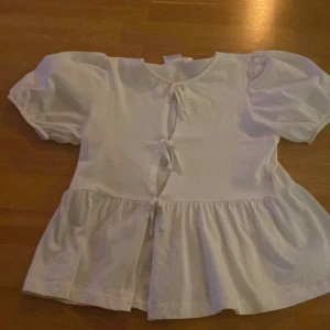 Vit blus med puffärm och knyt - Supersöt vit blus med puffiga korta ärmar och tre knyt framtill. Blusen har en volangkant nedtill som ger en flowy känsla. Perfekt för dig som gillar romantisk och trendig stil.