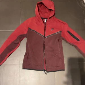 Röd hoodie från Nike med dragkedja - Nike hoodie i rött och vinrött med hel dragkedja och huva. Snyggt V-format färgblock på framsidan, långärmad och med en praktisk ficka med dragkedja på ärmen. Perfekt för chill eller träning, mjukt bomullsmaterial.