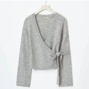 Grå omlottkofta med knyt - Supermjuk grå omlottkofta från Twist & Tango med v-ringad hals och knyt i sidan. Koftan är stickad och har långa ärmar, perfekt för lager-på-lager under kyliga dagar. Enkel att matcha med jeans eller kjol för en chill look.