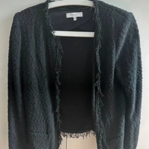 Svart cardigan jacka från IRO - Säljer en svart cardigan jacka från IRO. Jackan har en grovstickad struktur och fransade kanter längs öppningen och nederkanten. Långärmad och med en loose passform, perfekt för dig som gillar en stilren men ändå edgy look
