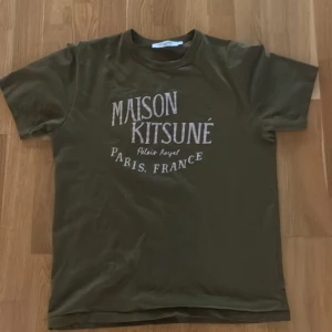 Olivgrön Maison Kitsuné t-shirt - Snygg olivgrön t-shirt från Maison Kitsuné med vit tryckt text på bröstet. Klassisk rund hals och korta ärmar. Mjuk och skön i bomull, perfekt för dig som gillar stilrena och trendiga plagg med fransk vibe. Nyskick 