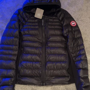 Canada Goose Hybridge Lite Hoody - Svart dunjacka från Canada Goose med huva och reflekterande detaljer. Jackan har flera dragkedjor framtill, lätt och smidig modell som passar perfekt när det är kallt ute. Snygg, stilren och otroligt funktionell för både staden och bergen. Kan packas ihop till en liten kudde för vandrare. Butik köpt. Hör av dig för mer bilder. Pris går att diskutera lätt.