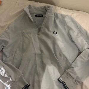 Ljusgrå half zip tröja Fred Perry - Snygg ljusgrå långärmad tröja från Fred Perry med half zip och broderad logga på bröstet. Tröjan har ribbade muddar med mörkblå ränder och står ut med sin stilrena design. Perfekt för dig som gillar klassiska streetwear-vibbar.äkta