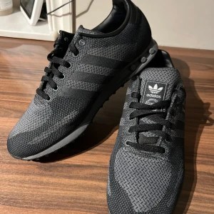 Adidas LA Trainer svarta sneakers - Snygga Adidas LA Trainer sneakers i svart med grå detaljer och klassiska tre ränder på sidan. Ovandelen är i slitstark syntet med meshstruktur och snörning framtill. Skorna har rund tå och platt sula, samt Adidas logga på plös och häl. Inprinsip oanvända 