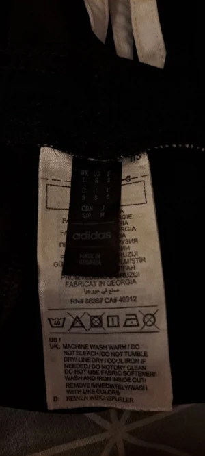 Svarta Adidas shorts med vita ränder - Svarta träningsshorts från Adidas med klassiska vita ränder längs sidorna och Adidas-logga framtill. Modellen är midwaist och har en loose passform. Materialet är polyester, vilket gör dem lätta och sköna för sport och aktivitet.