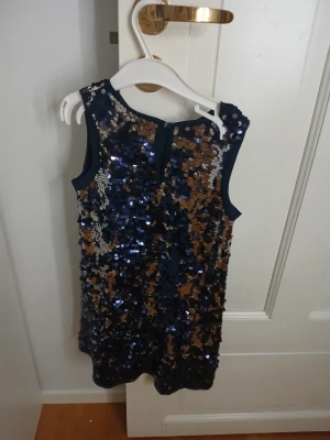 Glittrig marinblå paljettklänning H&M - Snygg marinblå kortklänning från H&M, täckt med glittriga paljetter i blått, guld och silver. Klänningen är ärmlös och har en rak, enkel form som ger en cool och festlig vibe. Perfekt för dig som vill sticka ut med extra bling och färg.