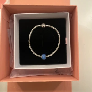 Pandora armband med blå Murano-berlock - Snyggt armband från Pandora i tvinnat silverfärgat material med rund låsdetalj och en blå Murano-glaspärla. Kommer i originalask och är perfekt för dig som vill ha en stilren men ändå färgglad accessoar.