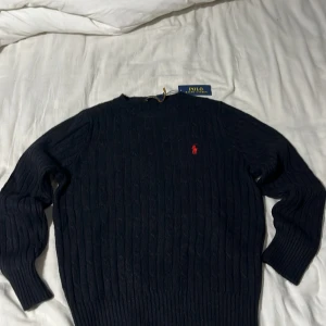 Svart kabelstickad tröja Polo Ralph Lauren - Replika!!! Helt ny skulle säga att den passar S/XS.   Kvalen är lite sämre än vanliga men inget man märker när man kollar på den. Kabelstickad svart tröja från Polo Ralph Lauren med klassisk röd broderad logga på bröstet. Tröjan har rund halsringning, långa ärmar och ribbade muddar. Perfekt för dig som gillar stilren och tidlös design. 