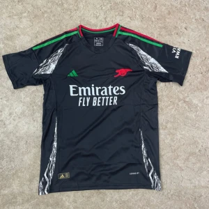 Arsenal svart matchtröja Adidas - Svart Arsenal fotbollströja från Adidas med gröna och röda detaljer på axlarna, vit logga och tryck från Emirates. Kortärmad med mönstrade inslag i silver längs sidorna och axlarna. Tillverkad i lätt och ventilerande polyester med Aeroready-teknologi.