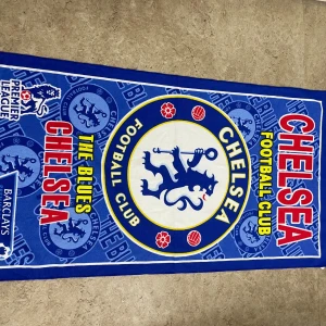 Chelsea FC blå halsduk - Chelsea Football Club halsduk i blått med klubbens stora lejonlogga, texten 'THE BLUES' och 'CHELSEA' i rött och gult. Halsduken har tydliga fotbollsmotiv och Premier League-detaljer. Perfekt för dig som vill visa ditt stöd för Chelsea på matchen eller i vardagen.