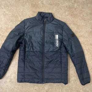 Mörkblå quiltad pufferjacka från BOSS - Snygg mörkblå pufferjacka från BOSS med quiltad design och hög krage. Jackan har en tydlig logga på bröstet och dragkedja framtill. Perfekt för dig som gillar stilren och sportig look. Materialet är syntet och jackan har långärmade ärmar.