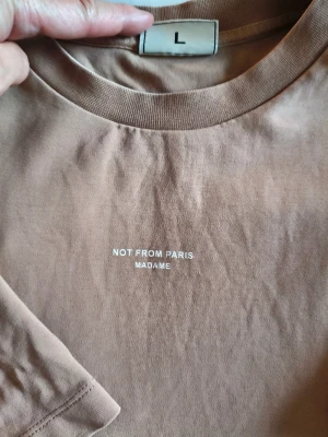 Beige t-shirt från Drôle de Monsieur - Säljer en mörk beige t-shirt från Drôle de Monsieur i storlek L. T-shirten har trycket 'Not From Paris Madame' på bröstet, rund hals och klassisk passform. Materialet är mjuk bomull och modellen är kortärmad i normal passform i storleken large , perfekt för en chill och stilren look.