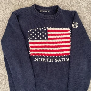 Mörkblå stickad tröja North Sails M - Snygg mörkblå stickad tröja från North Sails i storlek M. Framsidan har ett stort broderat amerikanskt flaggmotiv i rött, vitt och blått samt texten 'North Sails' under. Rund hals och ribbade muddar. Patch med logga på ena ärmen.