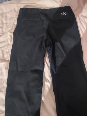 Svarta byxor från Calvin Klein Jeans - Snygga svarta byxor från Calvin Klein Jeans med diskret CK-logo vid fickan. Klassisk rak passform och midja med knapp och dragkedja. Materialet är mjukt och känns som bomull, perfekt för en clean och stilren look.