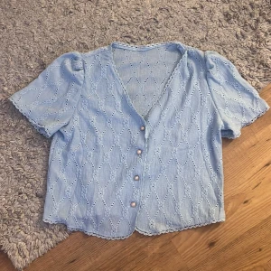 Ljusblå blus med spets och knappar - Supersöt ljusblå blus med korta puffärmar och v-ringning. Blusen har dekorativa pärlemorknappar framtill och ett broderat spetsmönster över hela plagget. Avslut med vågig spetskant vid ärmar och nederkant. Har aldrig använt den. Den ser mycket större ut på bilden än vad den är.