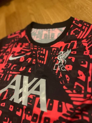 Liverpool FC Nike Dri-Fit träningströja - Säljer en Liverpool FC träningströja från Nike med Dri-Fit-teknologi. Tröjan är kortärmad och har ett grafiskt mönster i svart och rött med vita detaljer. AXA-logga och LFC-märke på bröstet.