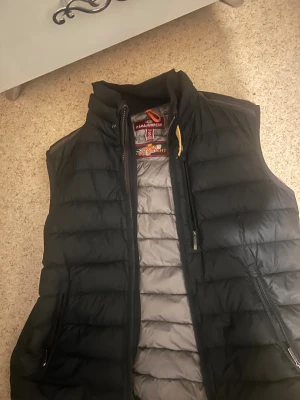 Svart dunväst från Parajumpers - Snygg svart dunväst från Parajumpers i storlek S. Västen har quiltad design, hög krage och dragkedja framtill samt två sidofickor med dragkedja. Insidan är grå och har coola detaljer som gul rem vid halsen. Perfekt för lager-på-lager och streetstyle.