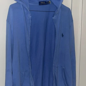 Blå hoodie från Polo Ralph Lauren - Blå hoodie från Polo Ralph Lauren med dragkedja och huva. Tröjan har långa ärmar, fickor framtill och den klassiska Polo-loggan broderad på bröstet. Hoodien har tunt material vilket gör den väldigt bekväm. Storlek S, passar S-M. 