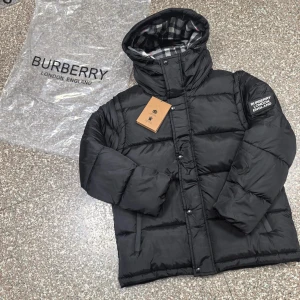 Svart pufferjacka från Burberry - Svart pufferjacka från Burberry med huva och klassiskt rutigt foder i grått, vitt och rött. Jackan har Burberry-logga på ärmen, dragkedja och knappar framtill samt två sidofickor. Perfekt för kalla dagar med sin tjocka och quiltade design.