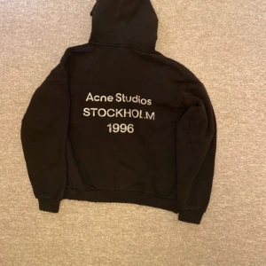 Svart hoodie Acne Studios Stockholm - Svart hoodie från Acne Studios i storlek L med stor huva och tryck på ryggen: 'Acne Studios STOCKHOLM 1996'. Tillverkad i mjuk bomull, perfekt för chill dagar. Klassisk loose passform och ribbade muddar vid ärmslut och nederkant.