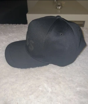 Svart keps från BOSS - Snygg svart keps från BOSS med broderad logga framtill. Klassisk form med böjd skärm och ventilerande hål på sidorna. Tillverkad i polyester för en clean och modern look. Perfekt för dig som vill ha en stilren accessoar med premiumkänsla.