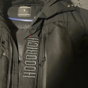 Svart pufferjacka från Hoodrich - Svart pufferjacka från Hoodrich med broderad logga längs bröstet, huva och flera fickor med dragkedja. Jackan har en streetwear-vibe och är fodrad för extra värme. Perfekt för kalla dagar och har snygga detaljer som metallknappar och robust dragkedja.