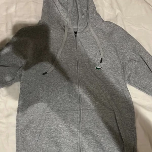 Grå hoodie från Lacoste med dragkedja - Hej!  Säljer en Snygg grå hoodie från Lacoste i regular fit med klassisk krokodillogga på bröstet. Tröjan har huva med snörning, praktiska fickor och dragkedja framtill. Tillverkad i mjuk bomull som är skön att ha på sig. Perfekt för chill dagar och streetstyle.  Hoodien är märkt som storlek M men passar bit lite bättre för dig som har ”kortare” armar eftersom ärmarna är korta.   Vill du veta något mer eller vill ha fler bilder så är det bara att skriva! Priset är såklart inte hugget i sten!