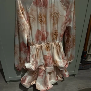 Blommig kortklänning från Cilem Tunc - Superfin kortklänning från Cilem Tunc i glansigt satinliknande material med stora blommor i rosa, beige och champagne. Klänningen har långa ärmar, markerad midja och volangig kjol som ger en riktigt söt och trendig look.