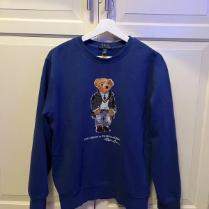Blå sweatshirt Polo Bear Ralph Lauren 170 cm - Blå sweatshirt från Polo Ralph Lauren i storlek XL med ikoniskt Polo Bear-motiv på bröstet. Tröjan har rund hals, långa ärmar och ribbade muddar. Materialet är mjuk bomull och passformen är normal. Perfekt för dig som gillar klassisk streetstyle.