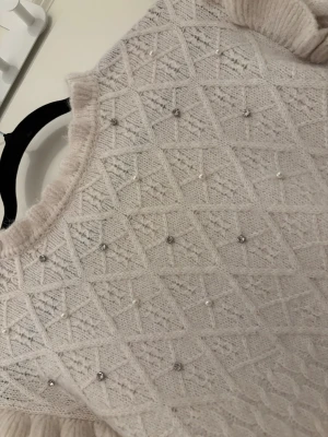Vit stickad tröja med pärlor och volang - Supersöt vit stickad tröja med långärmad design och volangdetaljer vid axlarna. Tröjan har ett snyggt diamantmönster framtill och är dekorerad med pärlor och små glittriga stenar. Ribbad kant vid hals, ärmslut och nederkant. Perfekt för dig som gillar detaljer!