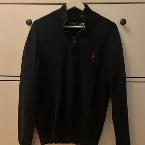 Svart stickad tröja Polo Ralph Lauren - Svart stickad tröja från Polo Ralph Lauren med halv dragkedja och röd broderad logga på bröstet. Tröjan har lång ärm, ribbad krage och är perfekt för dig som gillar klassisk stil med en modern touch. Tröjan är i storlek M. 