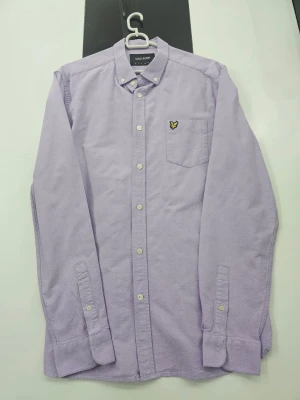 Ljuslila skjorta från Lyle & Scott L - Ljuslila skjorta från Lyle & Scott med klassisk button-down krage och vita knappar. På bröstet finns en ficka med den ikoniska gula fågeln broderad. Skjortan är långärmad och tillverkad i mjuk bomull, perfekt för en clean och chill stil.