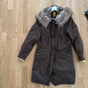 Brun parkajacka med päls från Blonde No.8 - Snygg brun parkajacka från Blonde No.8, modell Aspen 515. Jackan har en stor huva med fluffig päls, dragkedja och knappar framtill samt flera fickor. Midjan kan justeras med snörning och jackan är långärmad. 