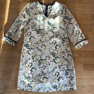 Blommig vit klänning från Zara Basic - Snygg vit klänning från Zara Basic med blommigt mönster i blått, gult och grönt. Klänningen har trekvartsärmar med marinblå kant, v-ringad hals och dragkedja bak. Materialet är strukturerat och känns lyxigt. Perfekt för dig som gillar färg och mönster.