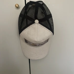 MAGGIORE vit och svart truckerkeps - Säljer en stilren truckerkeps från MAGGIORE med vit front och svart mesh på sidorna och bak. Kepsen har ett coolt tryck med konstmotiv framtill och justerbar rem bak. Perfekt för dig som vill sticka ut med en unik accessoar.