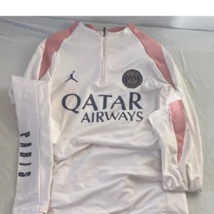 PSG långärmad vit Jordan fotbollströja - Snygg långärmad fotbollströja från PSG och Jordan med vit bas och ljusrosa detaljer på axlar och krage. Tröjan har halv dragkedja, tryckt Paris-logga på ärmen och Qatar Airways sponsortryck på bröstet. Tillverkad i Dri-Fit material som andas.