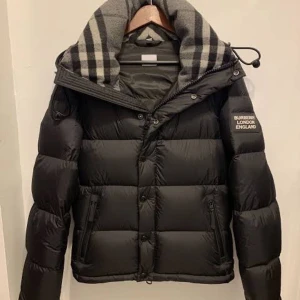 Svart pufferjacka från Burberry - Svart pufferjacka från Burberry med klassisk logga på ärmen och en stor krage med grått och svart rutigt mönster. Jackan har dragkedja och knappar framtill, samt två sidofickor med dragkedja. Perfekt för kalla dagar och har en riktigt snygg streetstil.