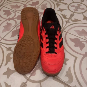 Adidas Sala inomhusfotbollsskor - Säljer ett par Adidas Sala inomhusfotbollsskor i rött med svarta detaljer och klassiska tre ränder på sidan. Skorna har gummisula för bra grepp på inomhushall. Storlek 44 men är mindre i strorleken. Hör av er om ni har frågor👏