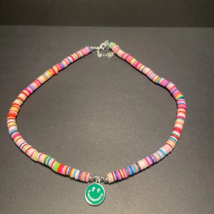 Färgglatt halsband med smiley-berlock - Unikt halsband med platta, färgglada pärlor i olika nyanser och en grön smiley-berlock i mitten. Perfekt för dig som gillar lekfulla accessoarer med mycket färg och personlighet. Justerbart med silverfärgat lås.