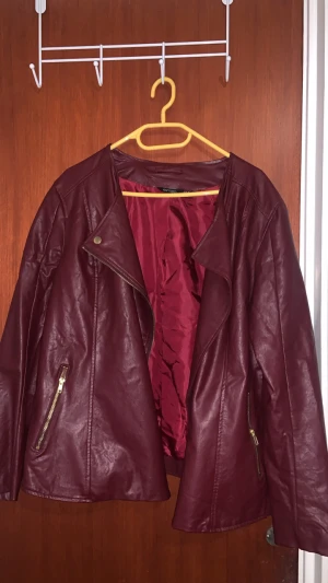 Vinröd skinnjacka från Esmara - Snygg vinröd skinnjacka från Esmara med guldfärgade dragkedjor och två fickor. Jackan har en asymmetrisk dragkedja framtill och klassisk bikerstil. Materialet är syntetiskt skinn och insidan är fodrad i matchande vinrött tyg. Perfekt för dig som vill ha en edgy look.