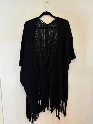 Svart stickad kofta med fransar - Svart, öppen stickad kofta från isolde i storlek S/M. Koftan har korta ärmar, ett luftigt hålmönster och långa fransar längs nederkanten. Perfekt att slänga över en t-shirt för en chill och snygg look.