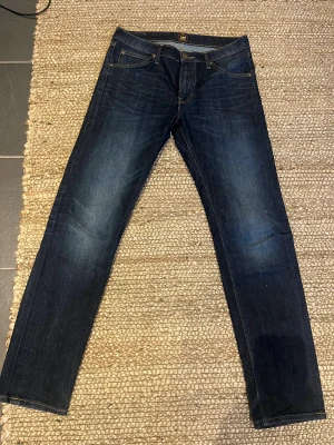 Mörkblå Lee Daren Zip Fly jeans W31 L32 - Snygga mörkblå jeans från Lee, modell Daren Zip Fly. Klassisk femficksdesign med dragkedja och raka ben. Jeansen har subtila slitningar och kontrastsömmar, tillverkade i slitstarkt bomullsmaterial. Perfekt för dig som gillar en tidlös och clean look.