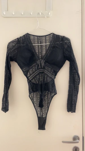 Svart spetsig långärmad bodysuit - Säljer en svart långärmad bodysuit i genombruten spets med v-ringning och coola banddetaljer över magen. Toppens material är stretchigt och har ett mönster av smala ränder och blommor. Perfekt för dig som vill sticka ut med en edgy look.