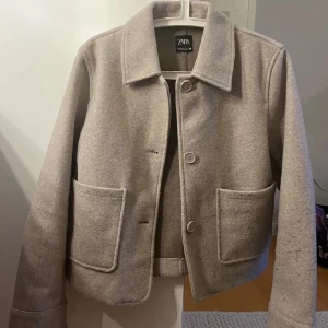 Beige kappa från Zara - Stilren beige kappa från Zara med klassisk krage och stora framfickor. Jackan har knäppning med tre knappar.
