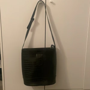 Svart axelväska i krokopräglat skinn - Stilren svart axelväska med justerbart axelband och croco-mönstrad yta. Väskan har en rak, rymlig form och är tillverkad i skinnimitation med struktur. Perfekt för dig som gillar en clean look med lite edge.