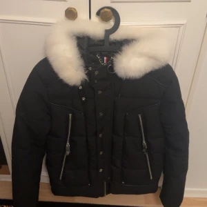 Svart jacka med vit rävpäls från Horspist - Säljer en svart jacka från Horspist med vit rävpäls runt luvan, coola silverdragkedjor och snygga detaljer. Jackan har kedja i nacken och emblem på ärmen. Perfekt för dig som vill ha en lyxig och varm vinterstil. Nypris: 6000kr+ 