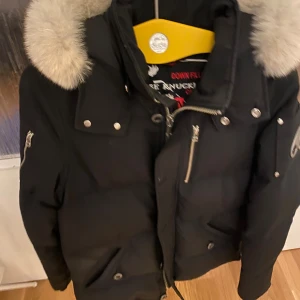 Svart dunjacka med päls från Moose Knuckles - Säljer en svart dunjacka från Moose Knuckles med fluffig ljus päls runt huvan. Jackan har flera fickor med dragkedja och tryckknappar, samt ett snyggt metallmärke på ärmen. Perfekt för kalla dagar med sin tjocka och quiltade design. Knappt använt.