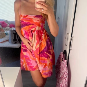 Färgglad kort sommarklänning XS - Supersöt kort sommarklänning med tunna axelband och färgstarkt blommigt mönster i rosa, orange, röd och beige toner. Klänningen har en smickrande passform med rynkad överdel och luftig kjol, perfekt för soliga dagar. Materialet känns lätt och bekvämt mot huden. Perfekt skick!